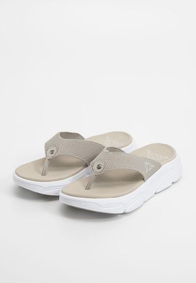 Des tongs beiges avec une lanière en tissu texturé, présentant un accent métallique circulaire et une semelle blanche épaisse avec un léger motif ondulé.