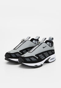 Paire de chaussures de sport grises et noires à enfiler, avec languettes, fermeture éclair et semelles blanches rembourrées, présentées sur fond blanc.